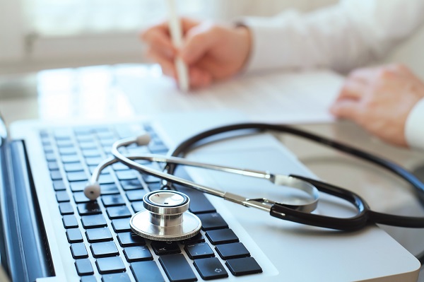 Stethoscope on a keyboard in forground with clinician writing in the background